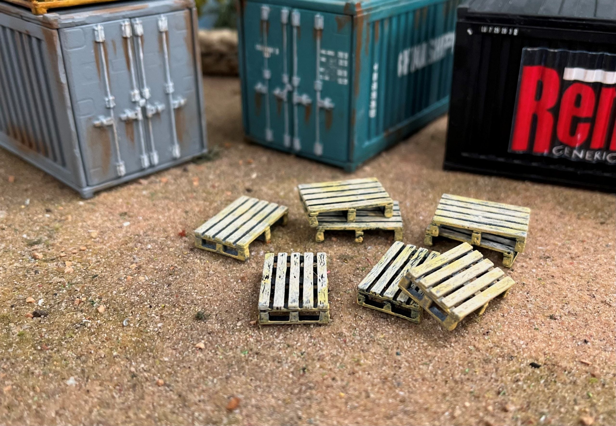 Painted 28mm scale Renedra hard plastic pallets scattered on a sandy yard in front of metal shipping containers.