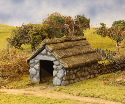 Fully assembled and painted 28mm scale Stone and Thatched Outbuilding shown in a rural wargaming diorama.