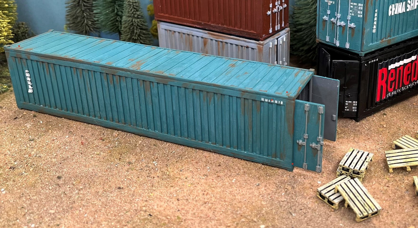 Fully assembled and weathered teal 40ft shipping container with open doors next to wooden pallets in a modern wargaming layout