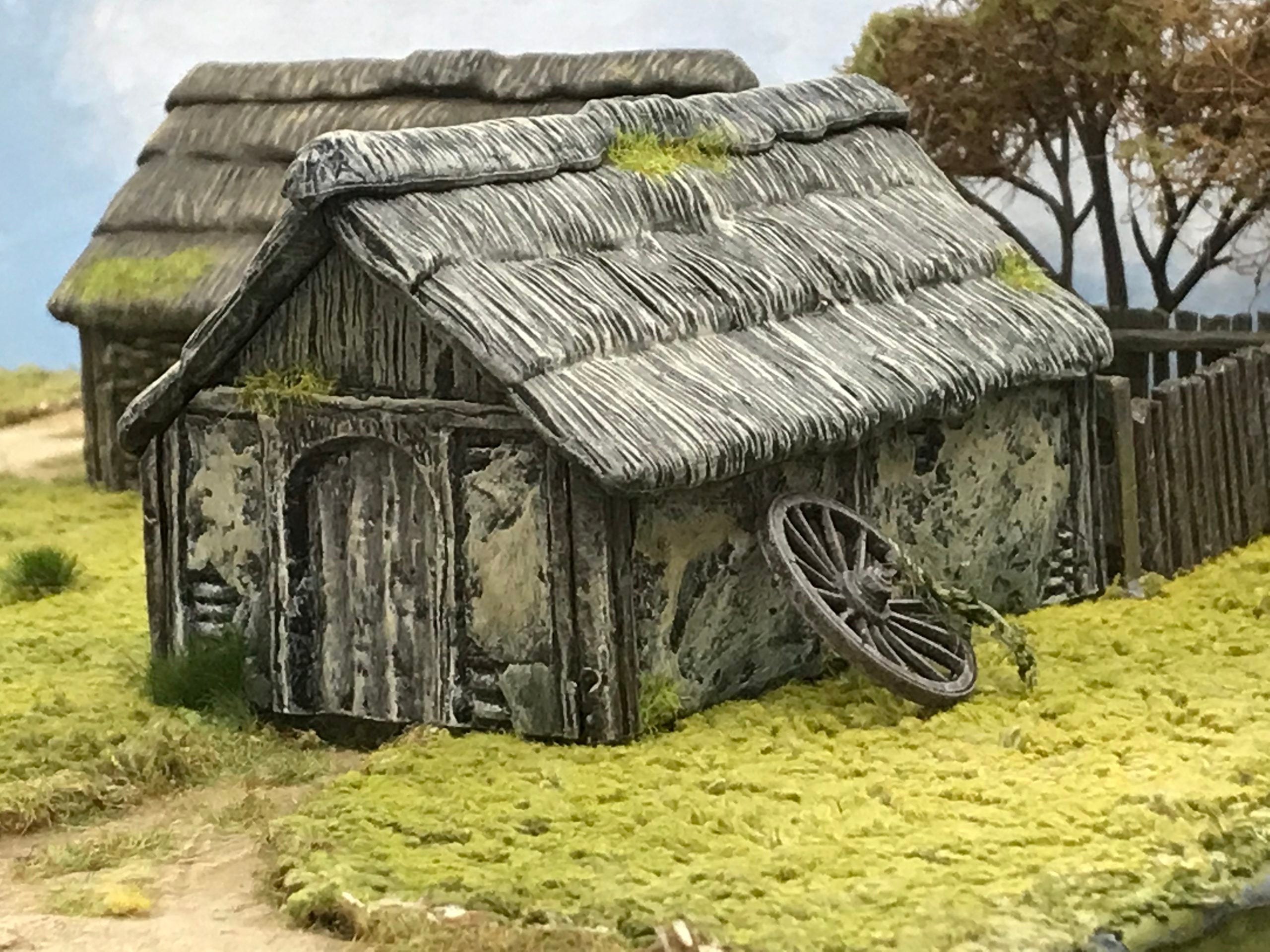 Fully assembled and painted 28mm scale Wattle and Daub Outbuilding featuring a thatched roof next to a wooden cart wheel accessory in a countryside display
